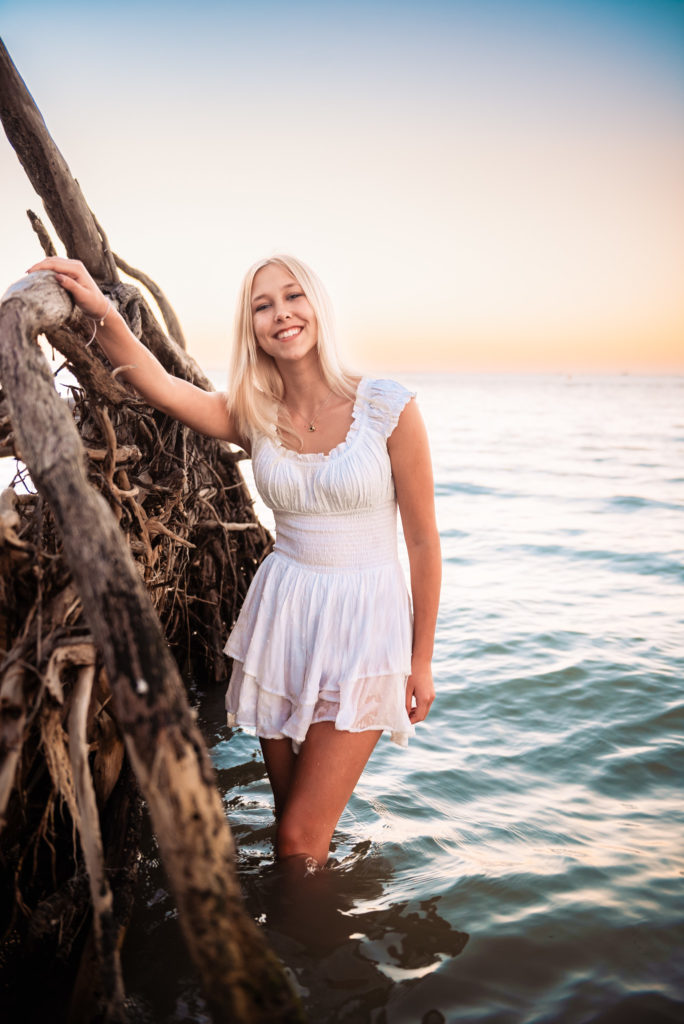 Girl in white romper posing with driftwood in the ocean on the beach.