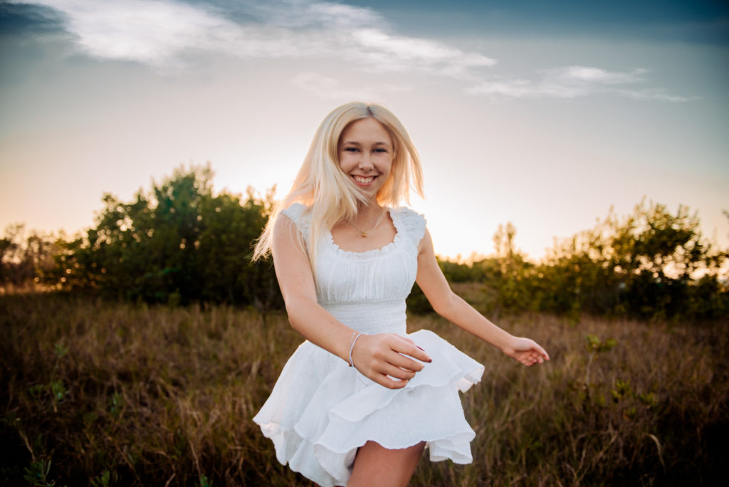 Graduating Senior Girl dancing in a field in a white romper. She is smiling and looking at the camera